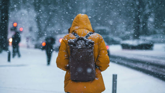 A person wearing a bright orange winter coat and carrying a snow-covered backpack walks through a snowy city street. Snow is falling heavily, and the background shows blurred figures and cars with headlights glowing through the storm.